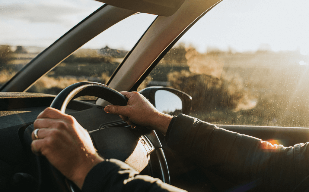 Imagem de uma pessoa dirigindo um carro, com as mãos no volante e um cenário iluminado pelo pôr do sol visível através do para-brisa.