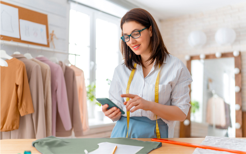 Uma mulher jovem, com cabelos castanhos e óculos, está sorrindo enquanto usa um smartphone. Ela veste uma camisa branca listrada e uma saia azul, com uma fita métrica amarela pendurada no pescoço. Ela está em um ateliê de costura, com roupas penduradas em cabides ao fundo e moldes de desenho fixados em um quadro na parede. Sobre a mesa à sua frente, há tecidos, uma régua longa laranja e papéis com moldes de costura. O ambiente é iluminado, com janelas grandes e um espelho decorado com luzes ao fundo para ilustrar o dia do consumidor