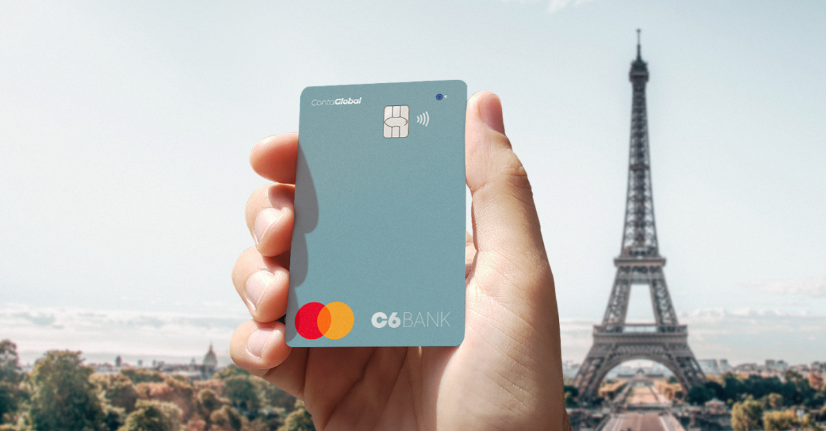 Imagem da mão de uma pessoa branca segurando o cartão azul claro da C6 Conta Global Euro na vertical com a Torre Eiffel ao fundo