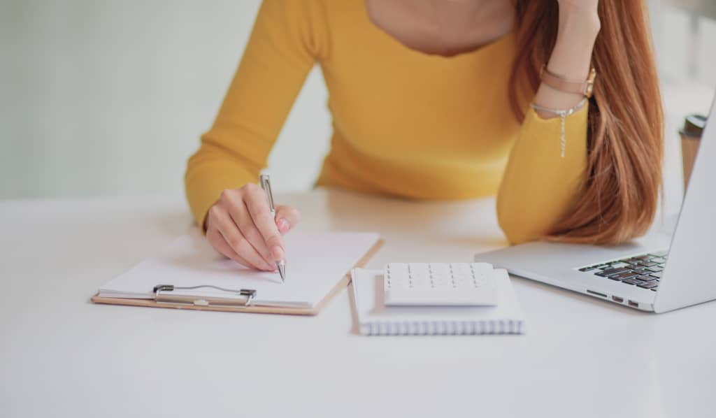 Mulher vestindo suéter amarelo escrevendo em uma prancheta sobre uma mesa branca. Ao lado, há um laptop, uma calculadora sobre um caderno e um copo de café, sugerindo um ambiente de trabalho ou estudo.