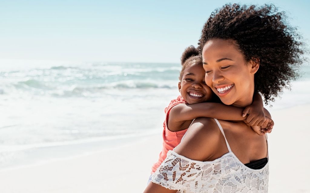 o que é mesada educativa mulher negra com uma criança abraça e apoiada em suas costas na praia